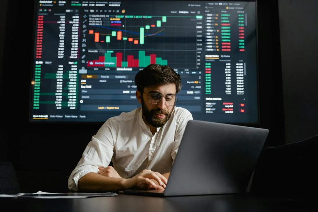 Man analyzing financial market charts on a laptop with a trading dashboard on a large screen