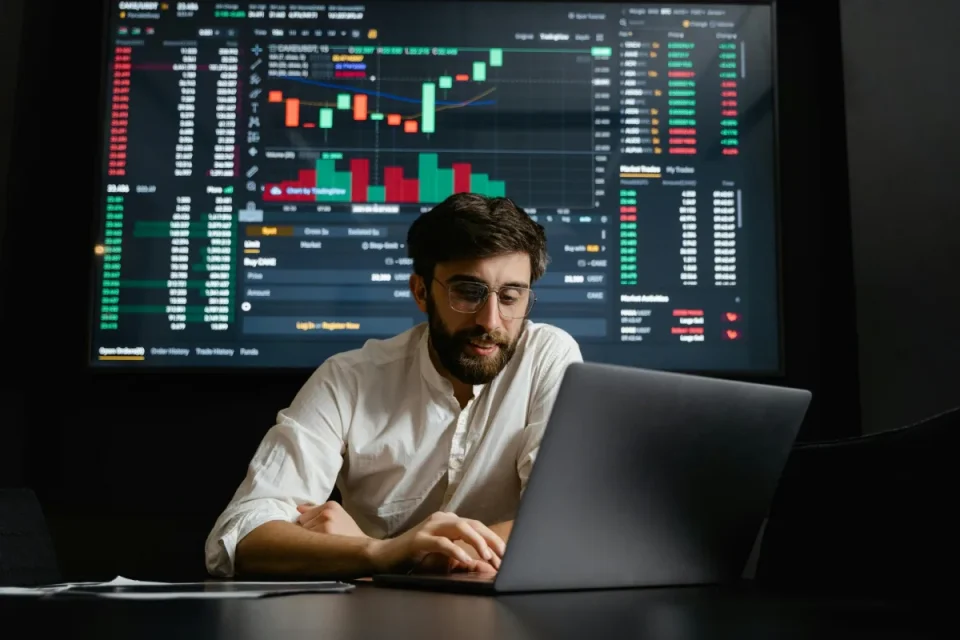 Man analyzing financial market charts on a laptop with a trading dashboard on a large screen