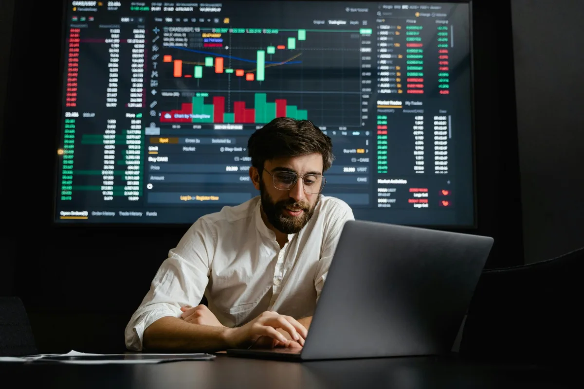Man analyzing financial market charts on a laptop with a trading dashboard on a large screen