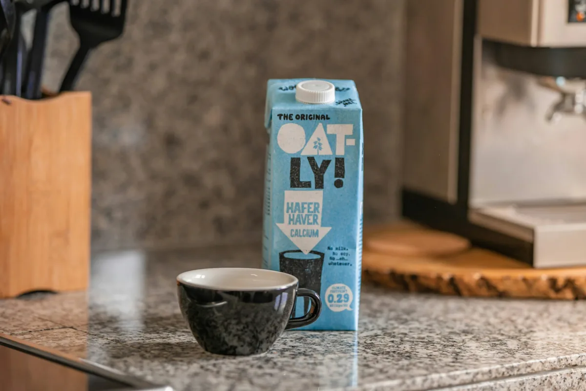 Oatly Barista oat drink next to a cup of coffee on a kitchen countertop