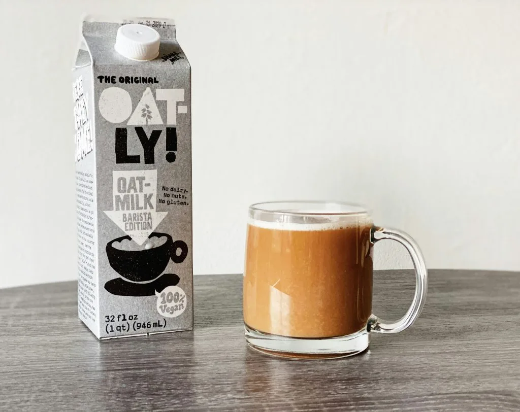 Oatly oat milk barista carton next to a glass mug of coffee on a table
