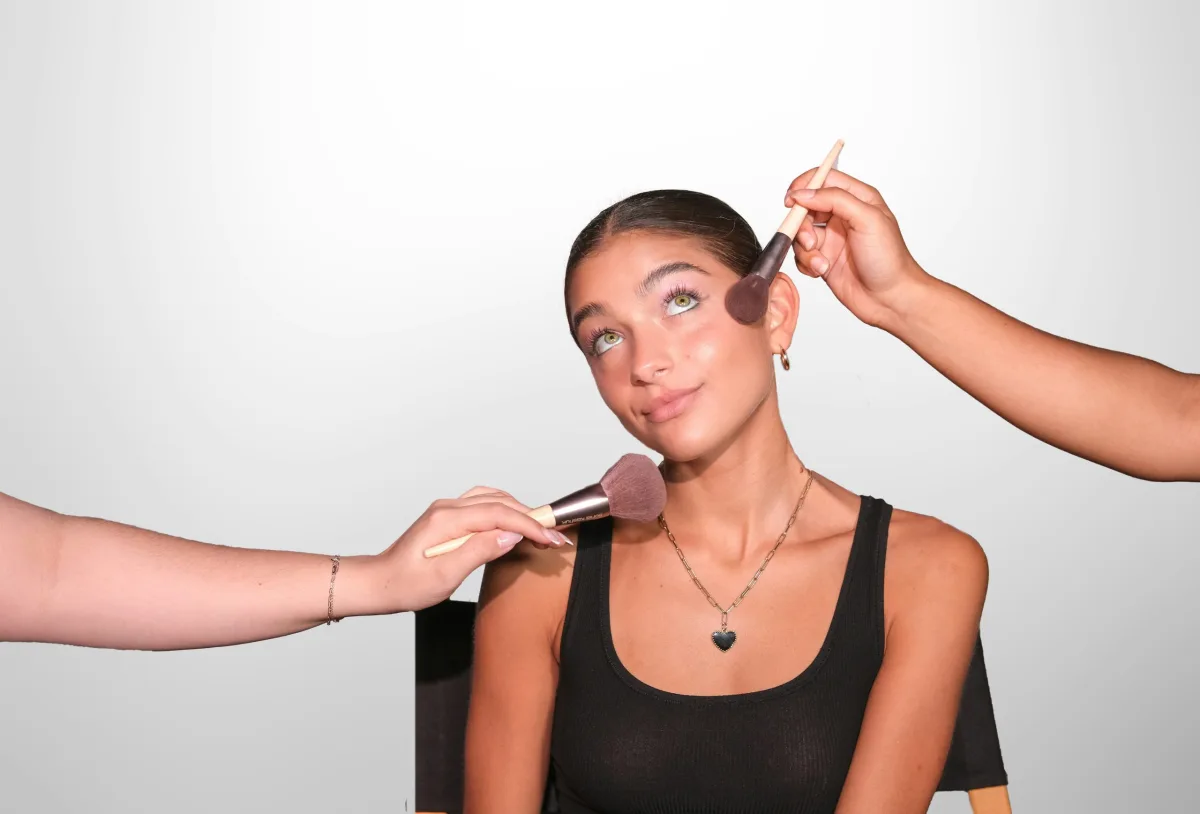 A woman sits on a chair while makeup artists apply makeup to her face with brushes; a makeup session against a light background.
