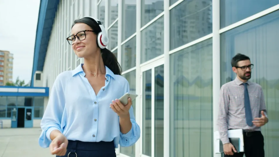 A woman in a blouse and headphones walks past a modern office building holding a smartphone; a man in a shirt and tie in the background.
