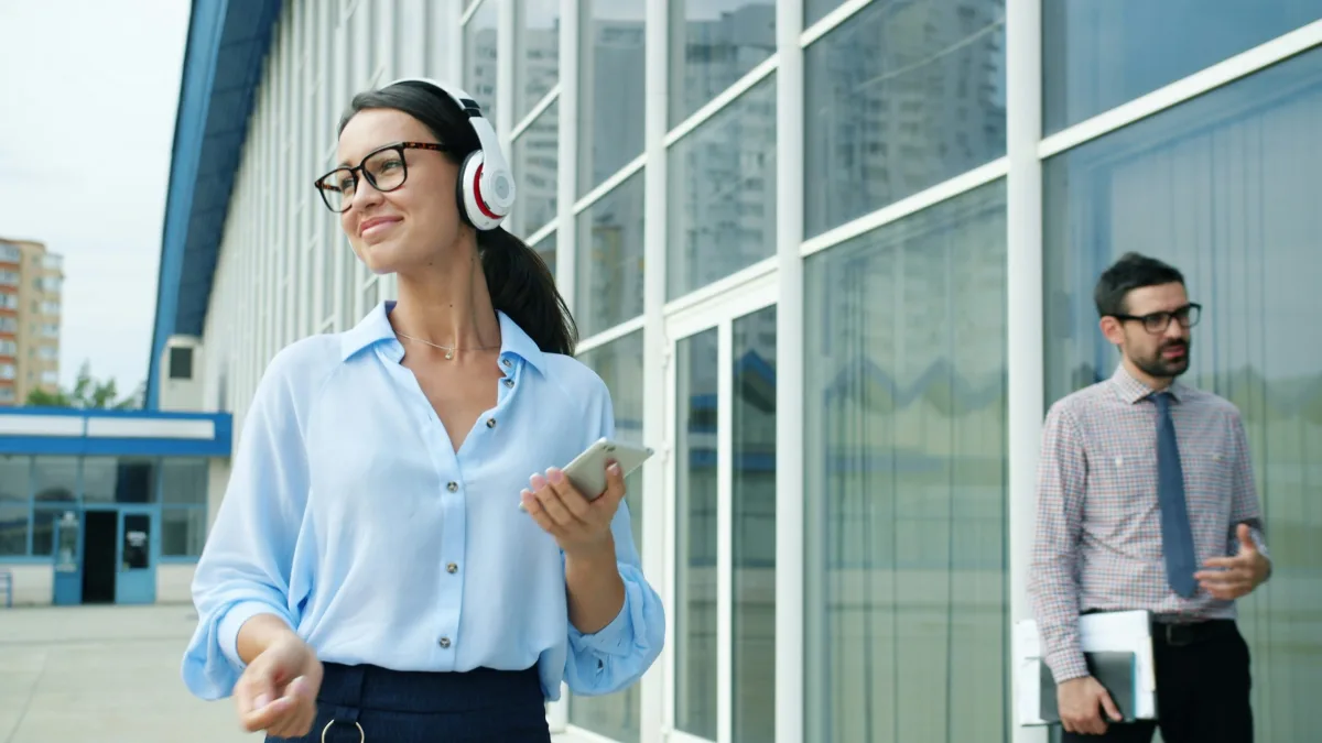 A woman in a blouse and headphones walks past a modern office building holding a smartphone; a man in a shirt and tie in the background.
