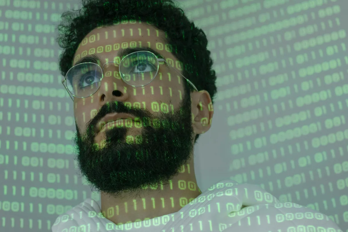Man with glasses and beard illuminated by green binary code projection on his face.