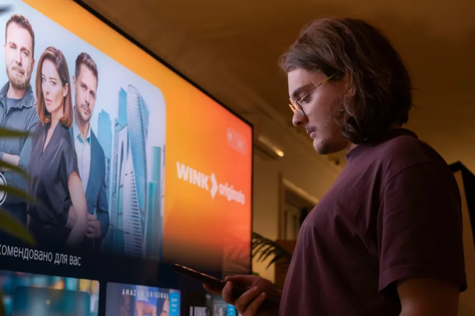Man using a phone while watching content on a large TV screen in a dimly lit room
