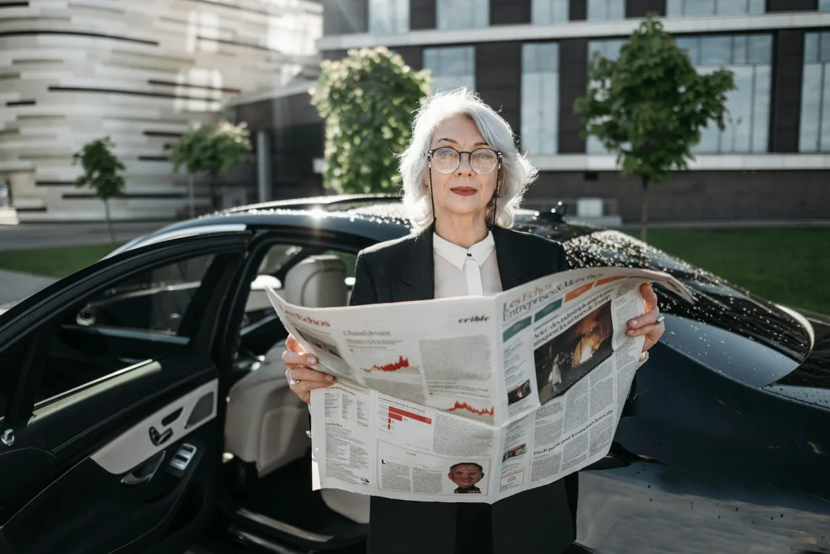 Senior businesswoman reading a financial newspaper beside a luxury car in a modern city setting