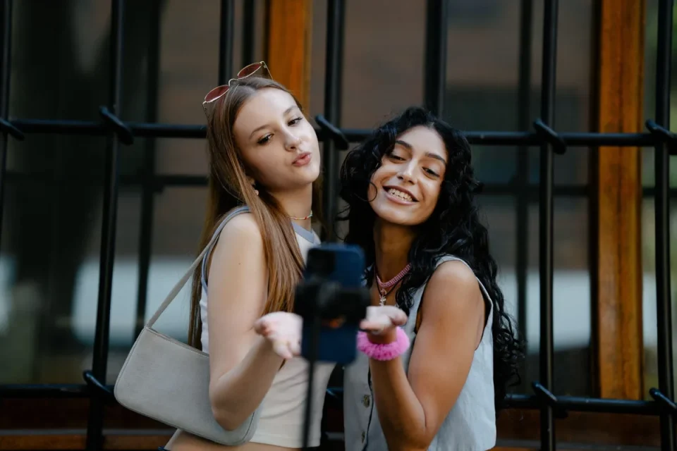 Two smiling young women posing for a selfie with a smartphone mounted on a selfie stick outdoors.