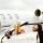 Woman in sunglasses and gold heels sitting in a white convertible parked next to a private jet on the tarmac, with her feet resting on the open window.