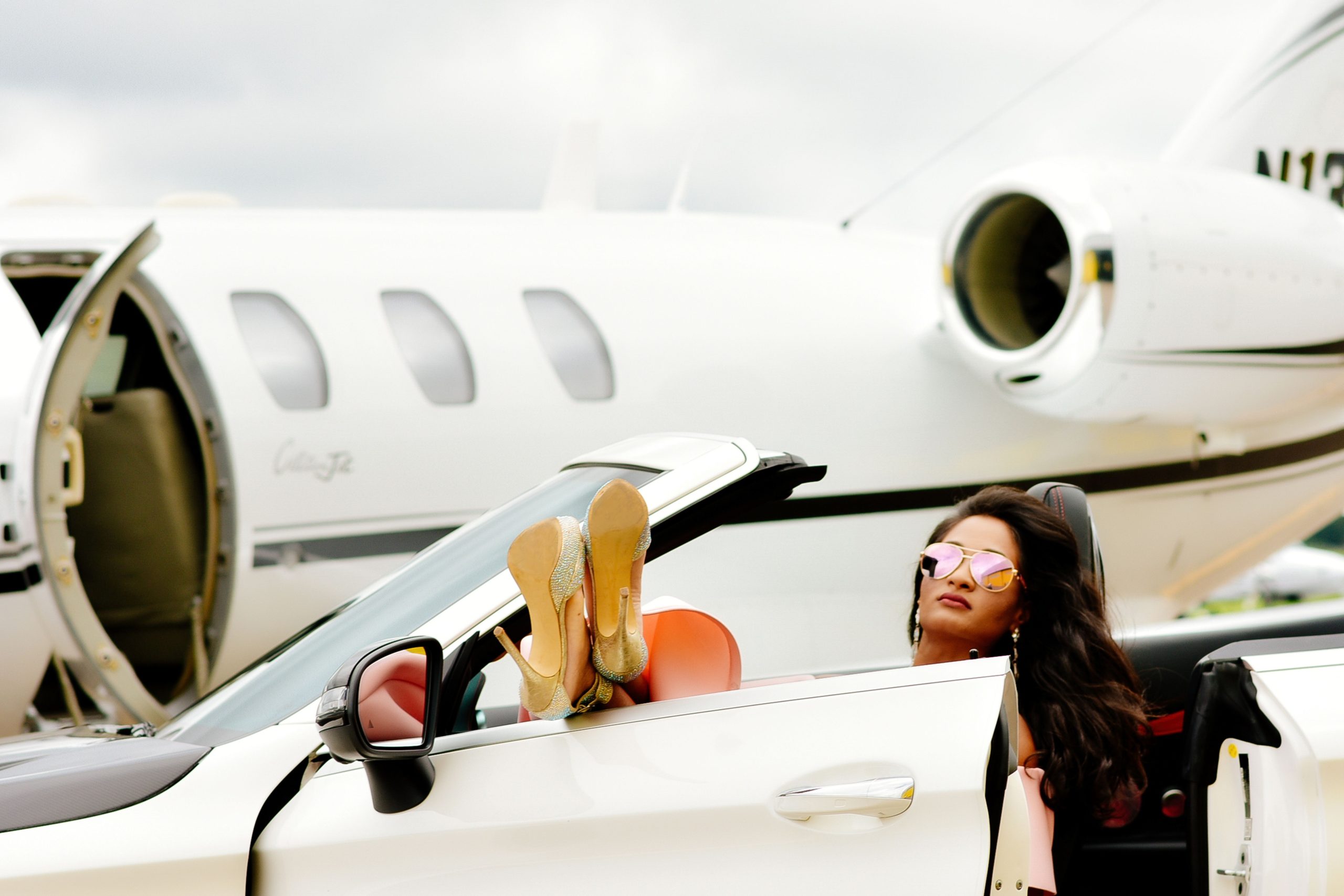 Woman in sunglasses and gold heels sitting in a white convertible parked next to a private jet on the tarmac, with her feet resting on the open window.