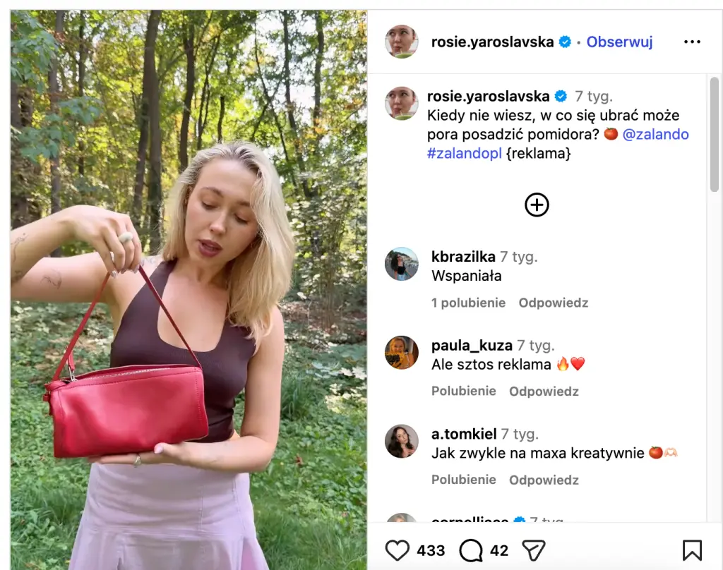 Young woman holding a red handbag while standing in a sunlit park surrounded by trees.